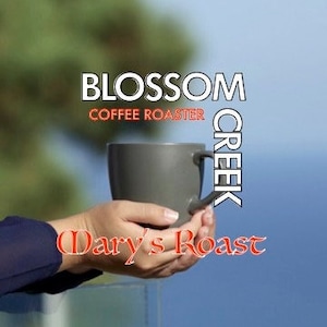 May include: A gray coffee mug held in hands, with the text "Blossom Creek Coffee Roaster" and "Dary's Roast" in the image. The background is a blurred view of greenery and a blue sky.