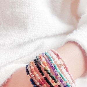 May include: A stack of colorful gemstone bracelets with gold accents. The bracelets are made of various stones, including amethyst, turquoise, and rose quartz.