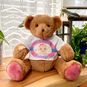 May include: A brown teddy bear wearing a white shirt with a pink and blue circle design. The circle has the text "Kailtyn Christine" and "Cartwright" inside.