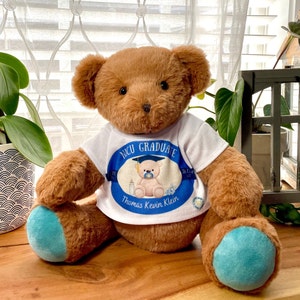 May include: A brown teddy bear wearing a white shirt with a blue circle that says "NICU GRADUATE" and "Thomas Kevin Klein". The bear has blue paw pads.