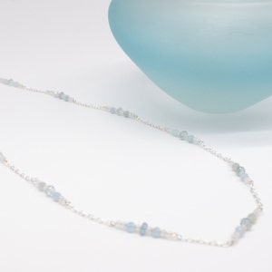 May include: A delicate necklace featuring small, light blue aquamarine beads strung on a fine silver chain. The necklace is displayed against a white background with a soft blue, abstract shape in the background. The beads vary in size and shade, creating a subtle, elegant design.