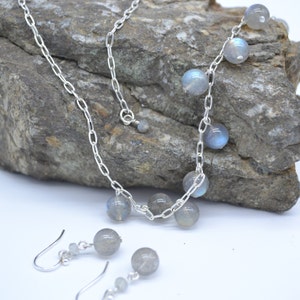 May include: A silver chain necklace with a silver chain drop earring set. The necklace has several round, light blue labradorite stones attached to it. The earrings have one round, light blue labradorite stone attached to each earring.