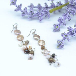 May include: A pair of dangle earrings with a mix of colors. Each earring features a series of flat, round, light-colored beads, with smaller, irregularly shaped beads in shades of brown, white, and pink dangling below. The earrings are set against a white background with purple flowers.
