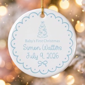 May include: A round white ceramic ornament with a scalloped light blue border. It features a light blue illustration of a Christmas tree, the text "Baby's First Christmas", "Simon Watters", and "July 9, 2026", finished with a gold hanging cord.