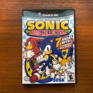 May include: A video game case for the Nintendo GameCube featuring Sonic the Hedgehog. The case is blue and black with a picture of Sonic, Tails, and Knuckles. The title of the game is "Sonic Mega Collection" and the text "7 Great Hits!" is also on the case.