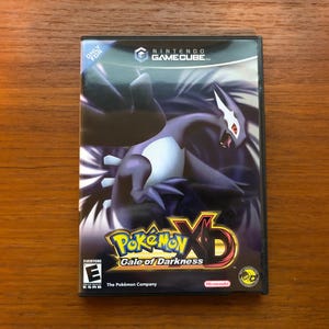 May include: A black Nintendo GameCube video game case for Pokémon XD: Gale of Darkness. The cover art features a purple and white Pokémon character with a red eye. The game title is in yellow and red text.