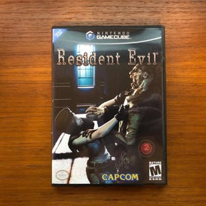 Puede incluir: Caja del videojuego Resident Evil para Nintendo GameCube. La portada presenta una escena dramática con dos figuras en un entorno oscuro. El título del juego se muestra de forma destacada, junto con el logotipo de Capcom y una clasificación.