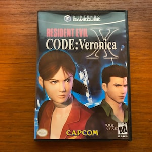 Könnte beinhalten: Resident Evil Code: Veronica X für Nintendo GameCube. Das Spielcover zeigt eine Frau und einen Mann mit dem Spieltitel in großen weißen Buchstaben. Das Capcom-Logo befindet sich unten. Freigegeben ab 17 Jahren.