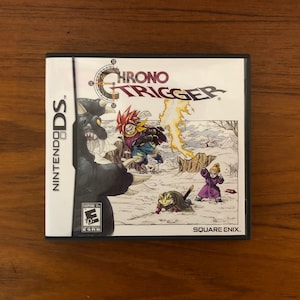 May include: A Nintendo DS game cartridge for Chrono Trigger. The game features a black and white illustration of characters from the game on a snowy background. The game's title is printed in red and yellow on a white background. The game is rated E for Everyone by the ESRB.