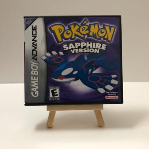 May include: A Game Boy Advance video game cartridge for Pokémon Sapphire Version. The game case is black with a purple background and features a blue and white creature. The game is displayed on a small wooden easel.