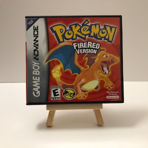 May include: A Game Boy Advance video game cartridge case for Pokémon FireRed Version. The case is red with the game title in yellow and a Charizard illustration. The case is displayed on a small wooden easel.