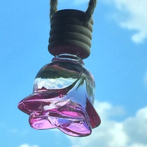 Könnte beinhalten: Ein rosa gläserner, rosenförmiger Lufterfrischer für das Auto, der an einer Schnur hängt. Der Lufterfrischer ist mit einer klaren Flüssigkeit gefüllt und hat einen Holzdeckel.