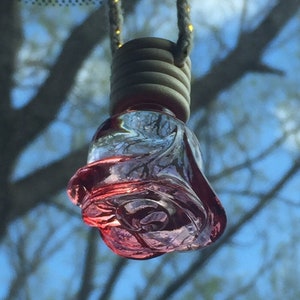 Op de afbeelding: Een roze glazen roosvormige autoluchtverfrisser die aan een touwtje hangt. De luchtverfrisser is van helder glas met een roze vloeistof erin.