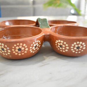 May include: A terracotta serving dish with four connected bowls. Each bowl is decorated with white and black floral patterns. A green handle connects the bowls. Suitable for serving snacks or dips.
