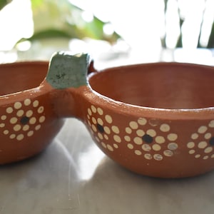 Könnte beinhalten: Ein terrakottafarbenes Serviergeschirr mit zwei Schalen und einem Verbindungsgriff. Die Schalen sind mit weißen und schwarzen Blumenmustern verziert. Der Griff ist hellblau-grün lackiert. Das Geschirr wird wahrscheinlich zum Servieren von Dips oder Snacks verwendet.