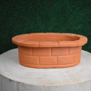 Könnte beinhalten: Ein ovaler Blumentopf in Terrakotta-Farbe mit einem ziegelartigen Muster an den Seiten. Er steht auf einer weißen, runden Holzoberfläche vor einem dunkelgrünen Hintergrund. Der Topf hat einen Durchmesser von etwa 20 cm und ist 10 cm hoch.