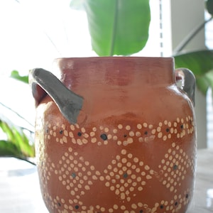 Pode incluir: Um vaso de cerâmica cor de terracota com boca larga e duas alças cinzentas. O vaso é decorado com um padrão de pontos brancos e pretos. O vaso é provavelmente usado para fins decorativos.