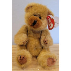 Peut inclure: Un ours en peluche brun doré avec une fourrure hirsute, un nez brun foncé et des yeux sombres. Une étiquette Ty rouge en forme de cœur est attachée à l'oreille, avec le texte blanc ty et la mention As Honey sur la partie blanche de l'étiquette.
