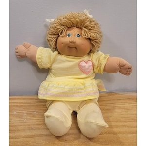 Pode incluir: Uma boneca de pano com pele bronzeada, olhos verdes e cabelo de lã loira decorado com fitas brancas. A boneca veste um vestido amarelo pálido de manga curta com um coração rosa aplicado no peito e listras na bainha.