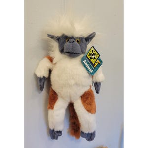 May include: A plush toy resembling a cotton-top tamarin monkey with white fur on the head and chest, grey face and limbs, and brown patches on the legs and tail. A diamond-shaped tag attached to the chest reads Make Noise! Animal.