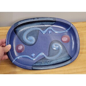 May include: An oval ceramic platter featuring a deep blue glaze with a speckled texture. The surface is decorated with two raised, light blue swirling lines and two small, circular reddish-purple accents positioned on opposite sides of the plate.