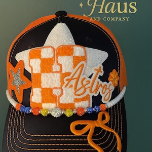 May include: A black and orange baseball cap with a white star and the word "Astros" in orange script. The cap features a checkered orange and white "H" and a beaded trim with colorful accents. The text "Hat Haus AND COMPANY" is in gold.