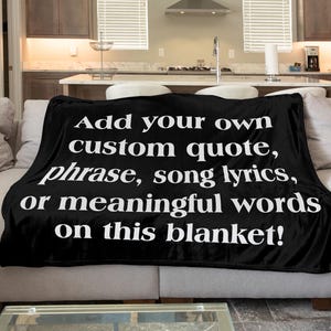 May include: A black rectangular blanket featuring white text that reads: Add your own custom quote, phrase, song lyrics, or meaningful words on this blanket! The blanket is draped over a light-colored sofa, displaying the text clearly in a serif font.