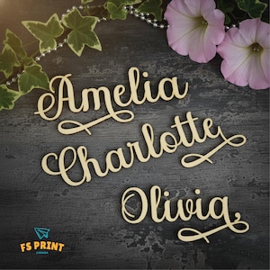 Könnte beinhalten: Holz-Namensschilder in einer Schreibschrift, die "Amelia", "Charlotte" und "Olivia" buchstabieren. Die Namen sind hellbraun und liegen auf einer dunkelgrauen Oberfläche, mit Blumen und Grünzeug im Hintergrund. Das Logo von FS Print Canada ist sichtbar.