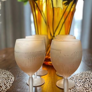 Schott Zwiesel Wine Glasses – Set of 6 – Peachy Grey Iridescent Six frosted wine glasses arranged on a wooden table with intricate lace-like placemats. A tall, amber vase with green branches adds elegance.