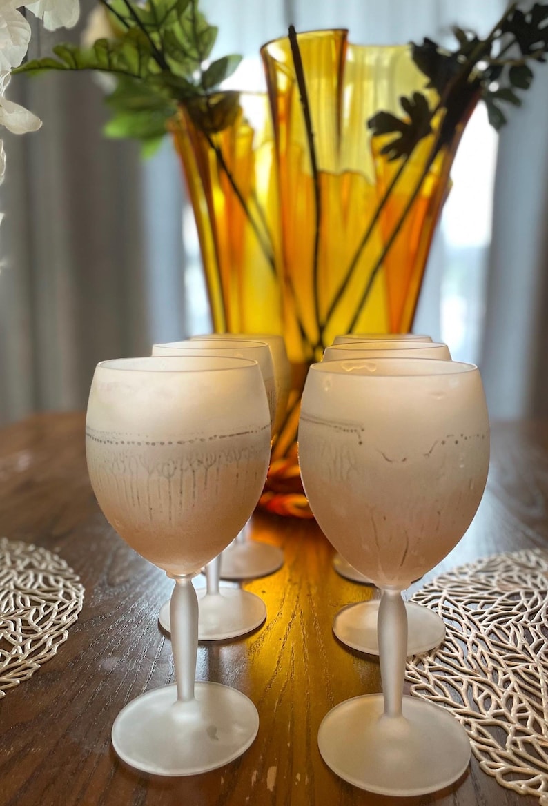 Schott Zwiesel Wine Glasses – Set of 6 – Peachy Grey Iridescent Six frosted wine glasses arranged on a wooden table with intricate lace-like placemats. A tall, amber vase with green branches adds elegance.