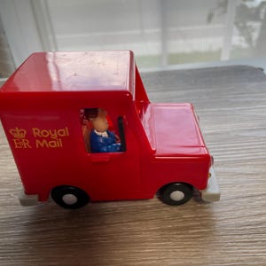 May include: A bright red toy mail delivery van featuring the Royal Mail logo and the ER emblem in gold lettering on the side. A small figurine wearing a blue uniform is visible in the driver seat. The vehicle has black wheels with white centres.