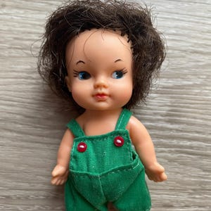 May include: A small doll with short, messy dark brown hair, blue eyes, and a light skin tone. The doll is wearing green fabric overalls featuring two small, round, bright red buttons on the front chest area, with bare arms and feet visible.