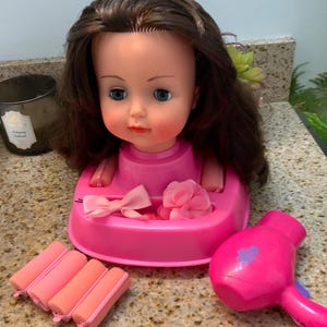 Puede incluir: Una cabeza de muñeca con cabello castaño y ojos azules, colocada sobre una base rosa con un lazo y una flor. Incluye rulos rosas y un secador de pelo rosa. Un juguete para peinados y juegos de belleza.