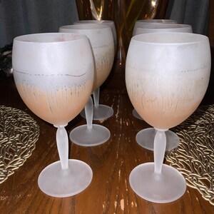 Schott Zwiesel Wine Glasses – Set of 6 – Peachy Grey Iridescent Six frosted wine glasses arranged on a wooden table with intricate lace-like placemats. A tall, amber vase with green branches adds elegance.