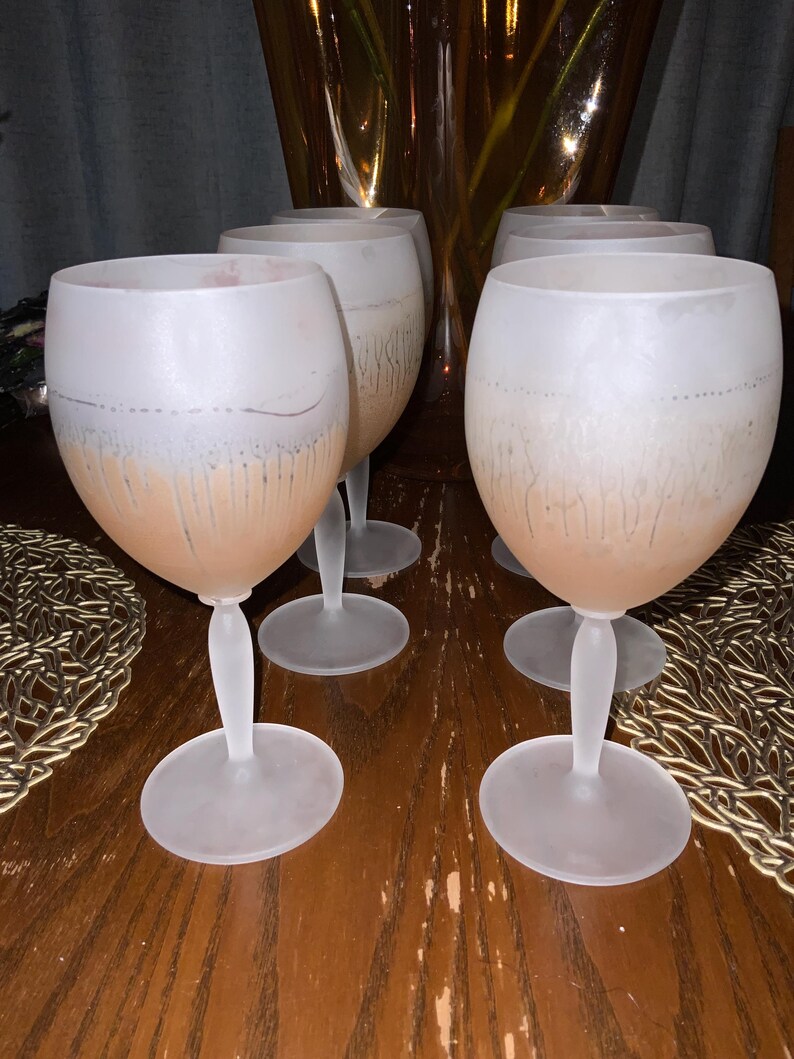 Schott Zwiesel Wine Glasses – Set of 6 – Peachy Grey Iridescent Six frosted wine glasses arranged on a wooden table with intricate lace-like placemats. A tall, amber vase with green branches adds elegance.