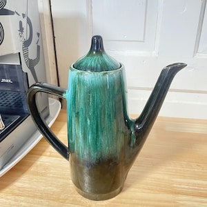 May include: A ceramic teapot with a tall, cylindrical body and a long, slender spout. The teapot is glazed in shades of green and brown, with a glossy finish. The handle and spout are black.