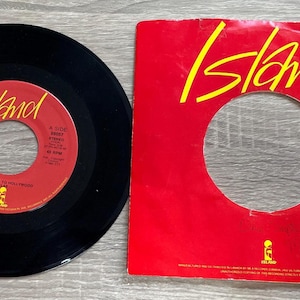 May include: A black vinyl record with a red label and a red sleeve. The label reads "Island" in yellow text. The sleeve also has the word "Island" in yellow. The record is a 45 RPM single.