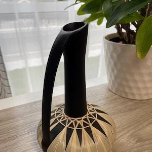 Puede incluir: Un jarrón negro con un asa curva y una base decorativa. La base presenta un patrón geométrico de material tejido beige y negro. El jarrón se exhibe sobre una superficie de madera de color claro, con una planta en el fondo.