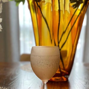 Schott Zwiesel Wine Glasses – Set of 6 – Peachy Grey Iridescent one frosted wine glass arranged on a wooden table with intricate lace-like placemats. A tall, amber vase with green branches adds elegance.