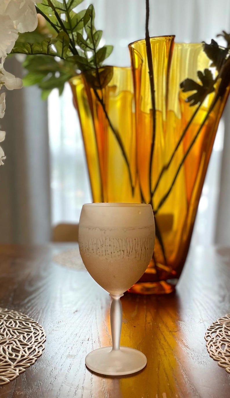 Schott Zwiesel Wine Glasses – Set of 6 – Peachy Grey Iridescent one frosted wine glass arranged on a wooden table with intricate lace-like placemats. A tall, amber vase with green branches adds elegance.