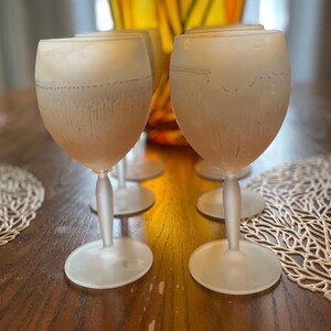 Schott Zwiesel Wine Glasses – Set of 6 – Peachy Grey Iridescent Six frosted wine glasses arranged on a wooden table with intricate lace-like placemats. A tall, amber vase with green branches adds elegance.
