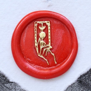 Puede incluir: Un sello de cera roja con un diseño dorado de una persona sentada en un trono. El sello está sobre un fondo blanco en forma de corazón.