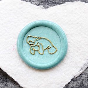 Peut inclure: Un sceau de cire bleu turquoise avec un contour doré d'un lamantin. Le sceau est sur un morceau de papier blanc en forme de cœur.