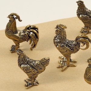 Puede incluir: Seis figuras de gallos de color bronce se disponen sobre una superficie beige. Los gallos tienen texturas de plumas detalladas y plumas de cola prominentes. Las figuras varían en tamaño y postura, creando una exhibición dinámica.
