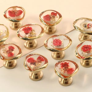 Puede incluir: Diez pomos de armario dorados con tapas de vidrio transparente y flores rojas prensadas en el interior. Los pomos son redondos y tienen un acabado liso y pulido.
