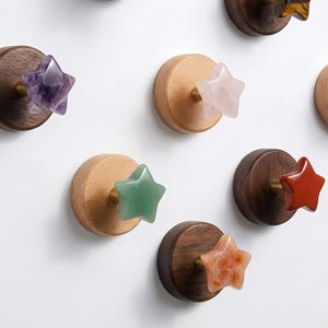 Op de afbeelding: Een verzameling decoratieve wandhaken met stervormige edelsteenaccenten. De haken zijn gemaakt van hout en messing, met edelstenen in verschillende kleuren, waaronder paars, groen, rood en oranje. De haken zijn op een witte muur gemonteerd.