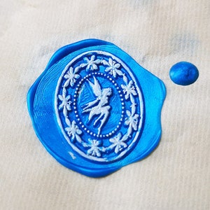 Peut inclure: Un sceau de cire bleu avec un motif blanc d'une fée avec les ailes déployées. Le sceau est sur une surface blanche.