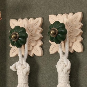 May include: Three decorative wall hooks. Each hook features a light wood carved floral design, a dark green knob, and a knotted white rope tassel. The hooks are mounted on a muted green wall.