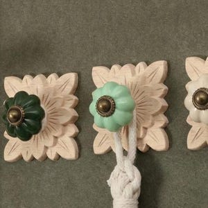 May include: Three decorative wall hooks with floral-shaped wooden backings. Each hook features a different colored knob: dark green, light green, and white. The knobs have a brass-colored center. A white tassel hangs from the center hook.