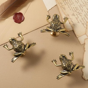 Könnte beinhalten: Drei messingfarbene Schrankknöpfe in Froschform mit ausgestreckten Gliedmaßen. Die Knöpfe haben eine detaillierte, strukturierte Oberfläche. Sie sind vor dem Hintergrund von Schreibwaren, darunter eine Feder, ein Umschlag und gealtertes Papier.